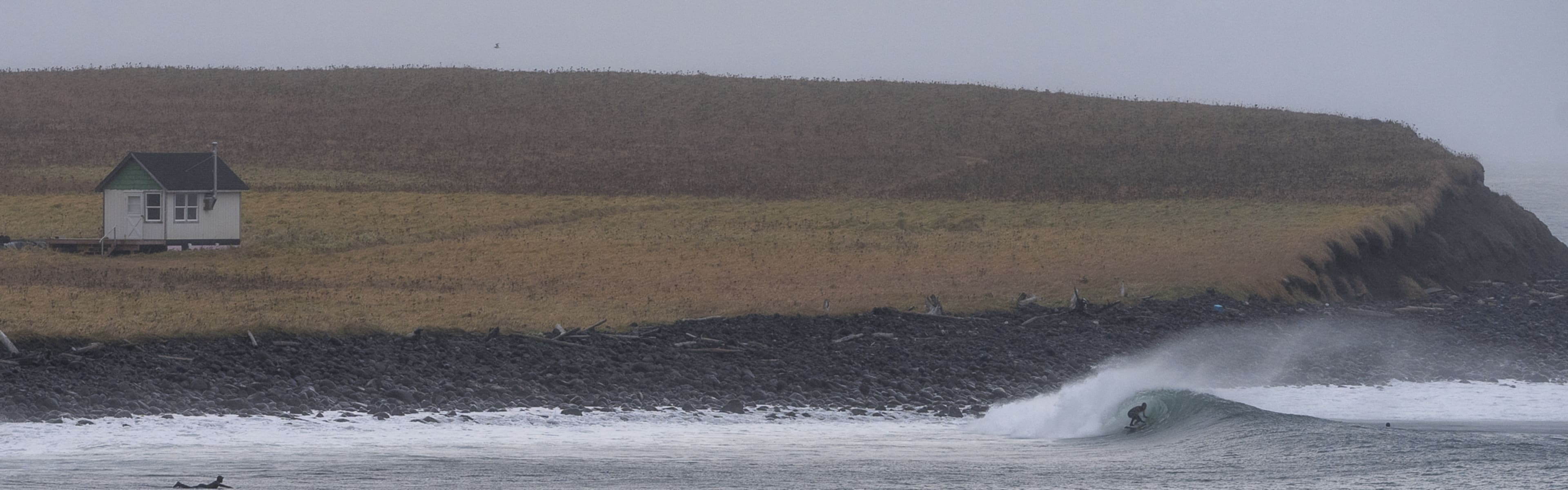 A left hand wave breaking in front of a desolate headland with a solitary home.